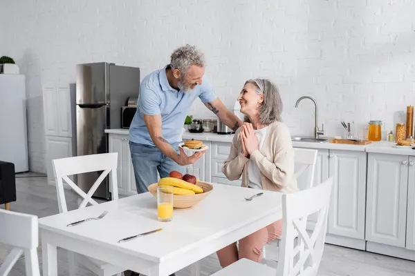 Man holding plate with pancakes while smiling wife showing please gesture in kitchen — Stock Photo