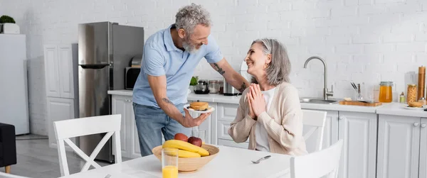 Femme souriante montrant s'il vous plaît geste près de mari mature avec des crêpes dans la cuisine, bannière — Photo de stock