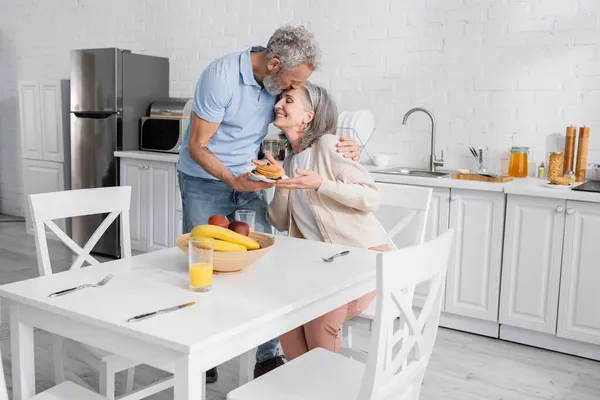 Hombre maduro besando esposa con panqueques cerca de jugo de naranja y frutas en la cocina - foto de stock