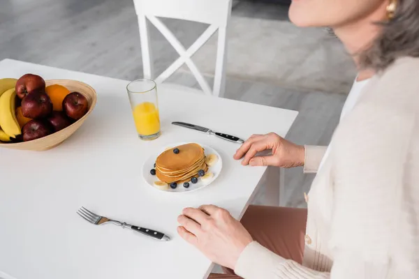 Cropped view of mature woman sitting near pancakes, orange juice and fruits in kitchen — Stock Photo