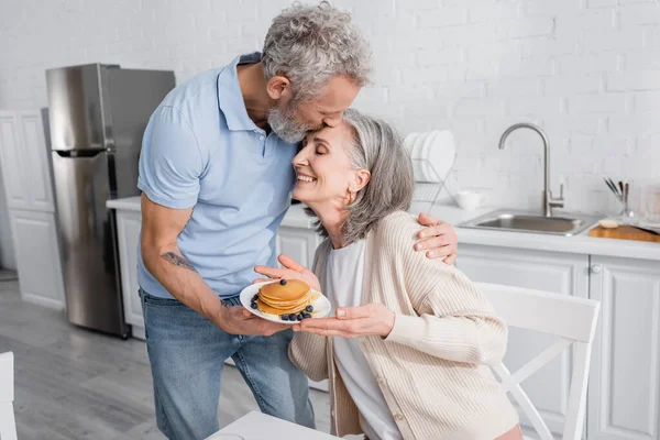 Hombre maduro besando esposa sonriente con sabrosos panqueques en la cocina - foto de stock