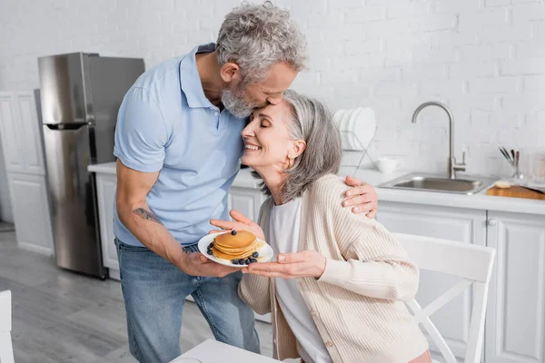 Mature man kissing smiling wife with tasty pancakes in kitchen — Stock Photo