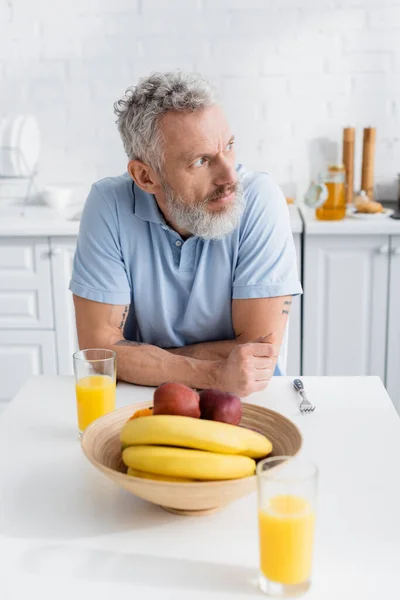 Зрелый татуированный мужчина смотрит в сторону фруктов и апельсинового сока на кухне — стоковое фото