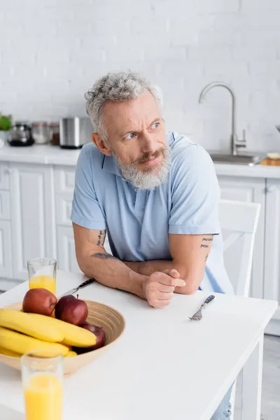 Мужчина средних лет сидит рядом с фруктами и апельсиновым соком на кухне — стоковое фото
