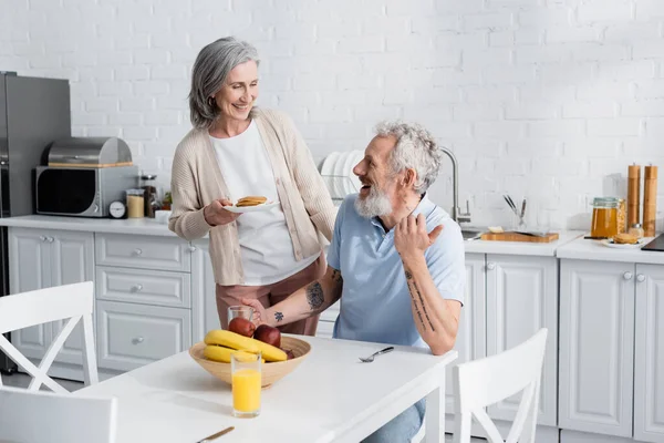 Mulher sorridente segurando panquecas perto de marido alegre e frutas na cozinha — Fotografia de Stock