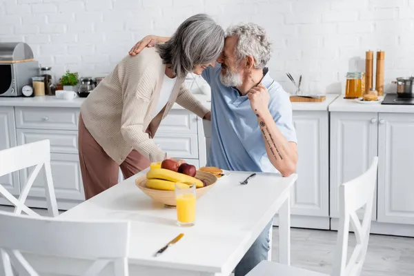 Smiling man hugging wife near breakfast and orange juice in kitchen — Stock Photo