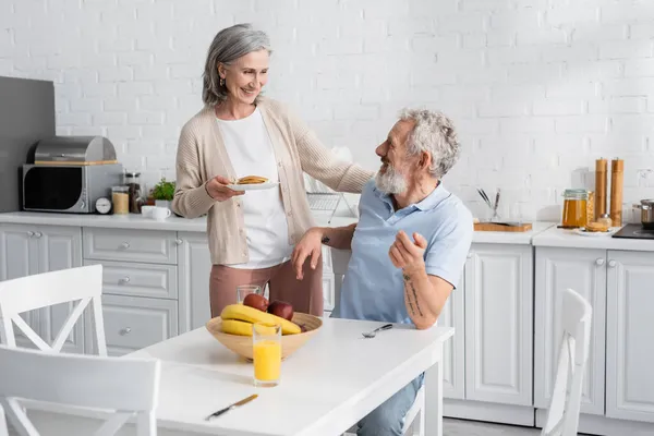 Smiling woman holding pancakes near husband and fruits in kitchen — Stock Photo