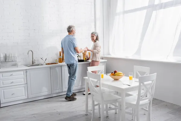 Pareja madura sonriente cogida de la mano mientras cocina en la cocina - foto de stock