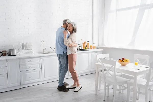 Positive woman hugging husband near fruits and orange juice in kitchen — Stock Photo