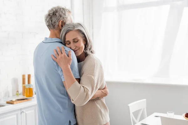 Жінка середнього віку з закритими очима, що обіймає чоловіка на кухні — стокове фото
