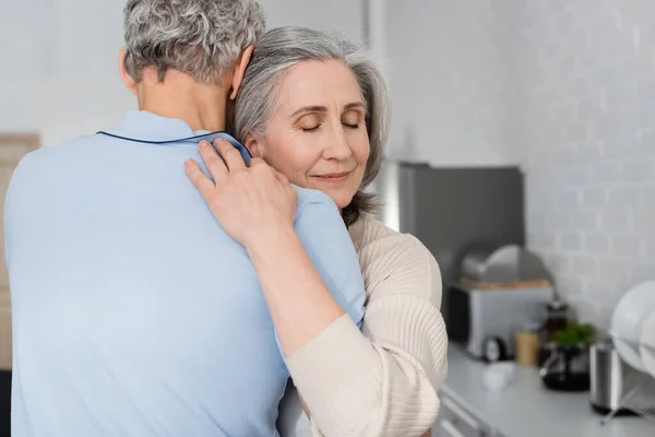 Donna matura con gli occhi chiusi che abbraccia il marito in cucina — Foto stock