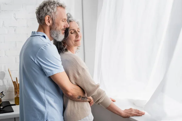 Reifer Mann umarmt Ehefrau in der Nähe von Vorhängen in Küche — Stockfoto