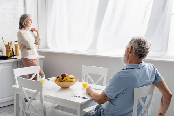 Homem maduro sentado perto de suco de laranja e esposa borrada na cozinha — Fotografia de Stock