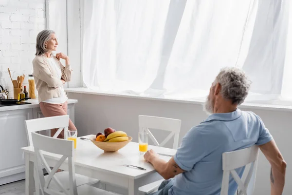 Mujer de mediana edad mirando a la ventana cerca del marido en la cocina - foto de stock