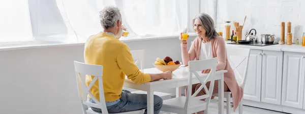 Lächelnde reife Frau hält Orangensaft neben Ehemann in der Küche, Banner — Stockfoto