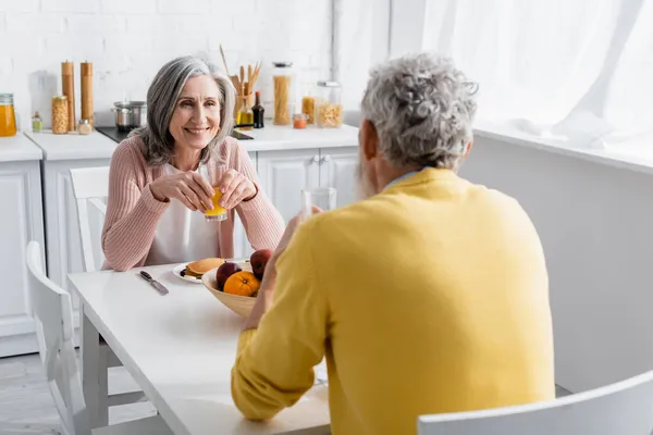 Donna positiva che tiene un bicchiere di succo d'arancia vicino a frutta e marito sfocato a casa — Foto stock