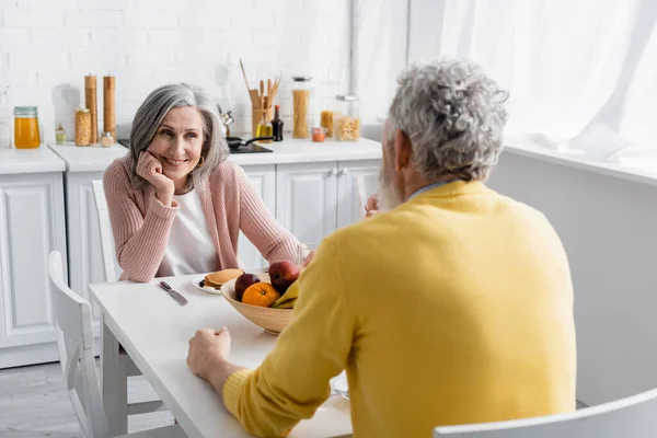 Mature woman looking at blurred husband near breakfast in kitchen — Stock Photo