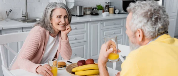 Frau mittleren Alters mit einem Glas Orangensaft in der Nähe des Frühstücks und verschwommenem Ehemann, Transparent — Stockfoto