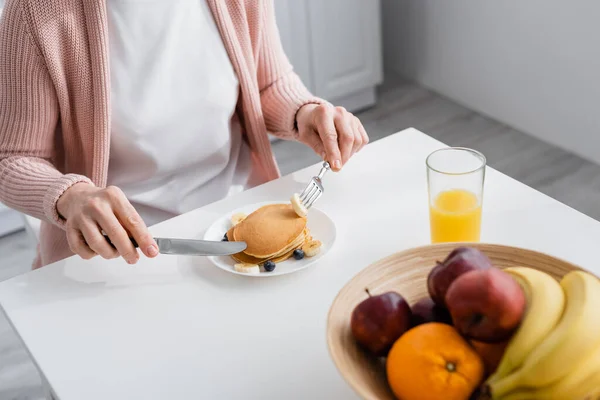 Cropped view of mature woman holding cutlery near pancakes and fruits at home — Stock Photo