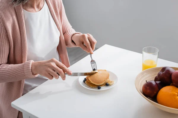 Ausgeschnittene Ansicht einer Frau, die Pfannkuchen in der Nähe von frischem Obst und Orangensaft in der Küche schneidet — Stockfoto