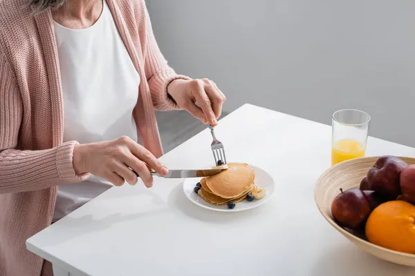 Cropped view of woman cutting pancakes near fresh fruits and orange juice in kitchen — Stock Photo