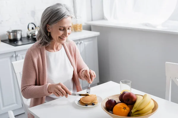 Mujer sonriente cortando panqueques cerca de frutas maduras en la cocina - foto de stock