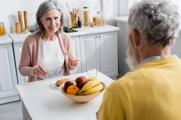 Cheerful mature woman looking at blurred husband near pancakes and fruits in kitchen — Stock Photo