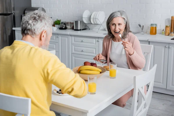 Зрелая женщина ест блинчики во время завтрака с мужем на кухне — стоковое фото