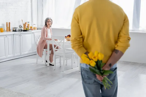 Smiling woman looking at blurred husband hiding flowers in kitchen — Stock Photo