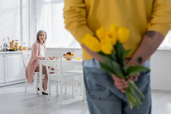 Middle aged woman looking at husband hiding tulips in kitchen — Stock Photo