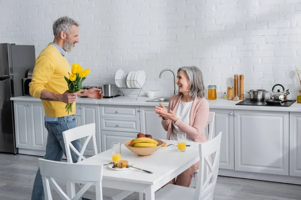 Vista lateral del hombre sosteniendo flores cerca de la esposa positiva y el desayuno en la cocina - foto de stock