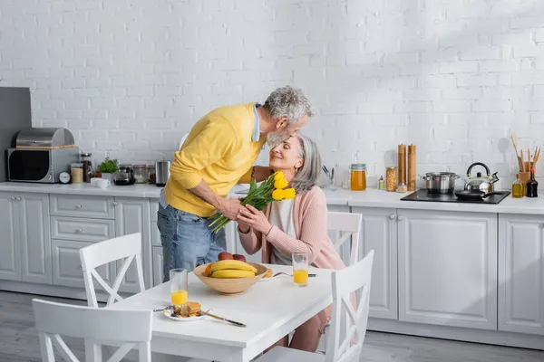 Мужчина с цветами целует жену во время завтрака на кухне — стоковое фото