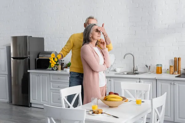 Homem segurando flores e cobrindo os olhos da esposa perto do café da manhã na cozinha — Fotografia de Stock