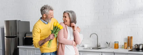 Smiling man hugging wife and holding yellow tulips in kitchen, banner. Translation: 