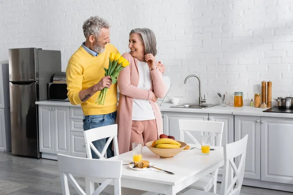 Hombre alegre sosteniendo tulipanes cerca de la esposa y desayuno en la cocina. Traducción: 