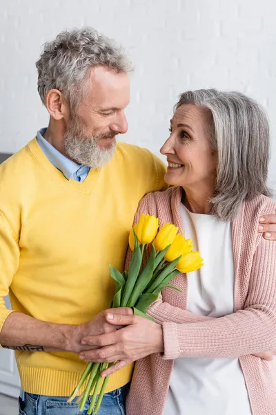 Mujer sonriente sosteniendo flores cerca de marido barbudo en casa - foto de stock