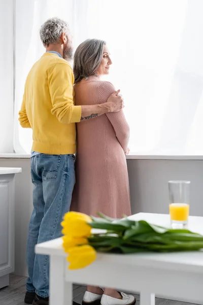 Чоловік обіймає дружину біля вікна і розмиті квіти на кухні — стокове фото