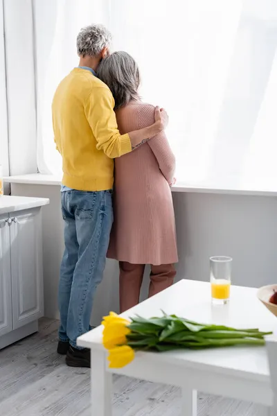 Vista posteriore della coppia matura che abbraccia vicino alla finestra e tulipani sfocati sul tavolo a casa — Foto stock