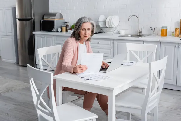 Mujer madura cuenta facturas cerca del ordenador portátil en la cocina - foto de stock