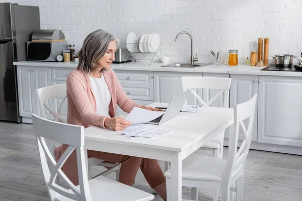 Mujer de mediana edad utilizando el ordenador portátil cerca de papeles con facturas en la cocina - foto de stock