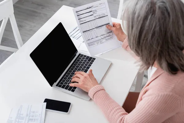 Ansicht der grauhaarigen Frau mit Laptop in der Nähe von Geldscheinen auf Tisch und Smartphone mit leerem Bildschirm — Stockfoto