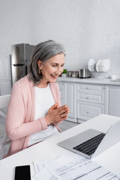 Mujer de pelo gris sonriendo y haciendo gesto de oración cerca de dispositivos y facturas en la cocina - foto de stock