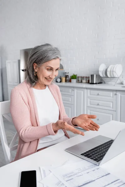 Mujer de mediana edad positiva apuntando a la computadora portátil durante la videollamada cerca de facturas en casa - foto de stock