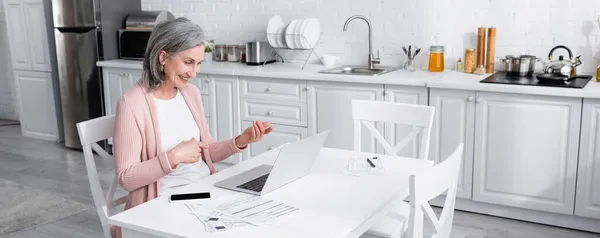 Femme mature gestuelle lors d'un appel vidéo sur un ordinateur portable près d'un smartphone et factures dans la cuisine, bannière — Photo de stock