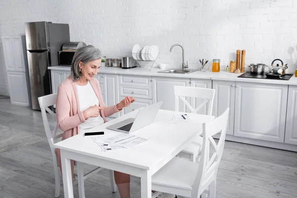Fröhliche reife Frau zeigt mit Fingern während Videoanruf auf Laptop neben Papier mit Scheinen in der Küche — Stockfoto