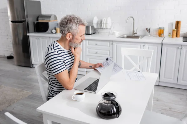 Vue latérale de l'homme barbu regardant les factures près de l'ordinateur portable et le café dans la cuisine — Photo de stock