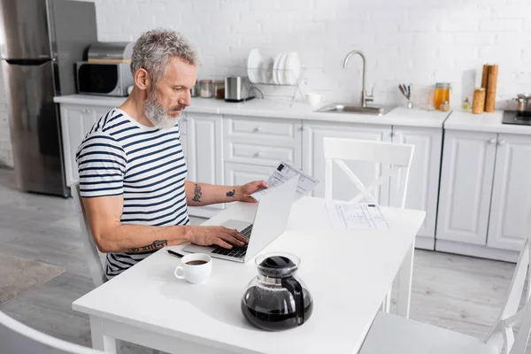 Homme utilisant un ordinateur portable tout en comptant les factures près du café à la maison. Traduction : 