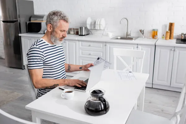 Mann mit Laptop beim Zählen von Rechnungen in der Nähe von Kaffee zu Hause. Übersetzung: 