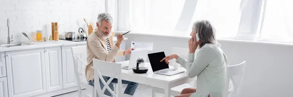 Hombre apuntando a la esposa hablando en el teléfono inteligente cerca de facturas y portátiles en la cocina, pancarta - foto de stock