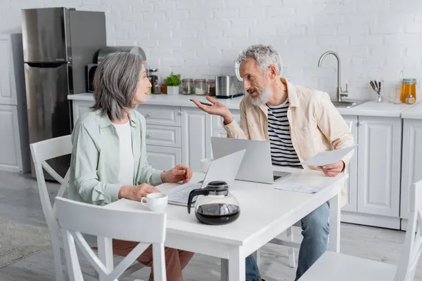 Ältere Paare reden in der Nähe von Rechnungen und Laptops in der Küche — Stockfoto